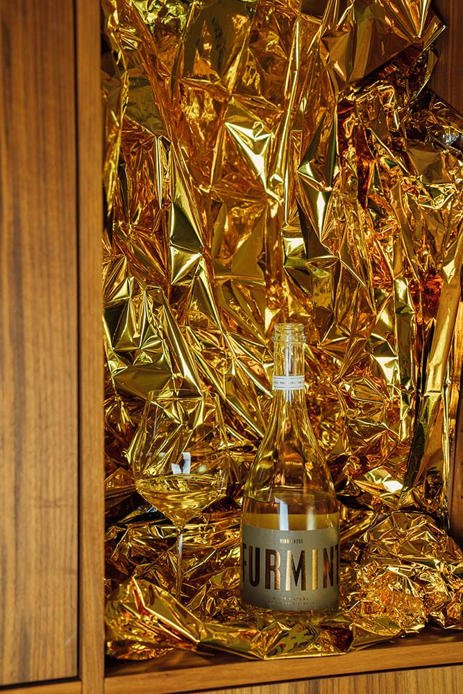 Eine Flasche Furmint Brut Nature 2013 von Vino Gross neben einem Glas. Im Hintergrund goldene Folie.